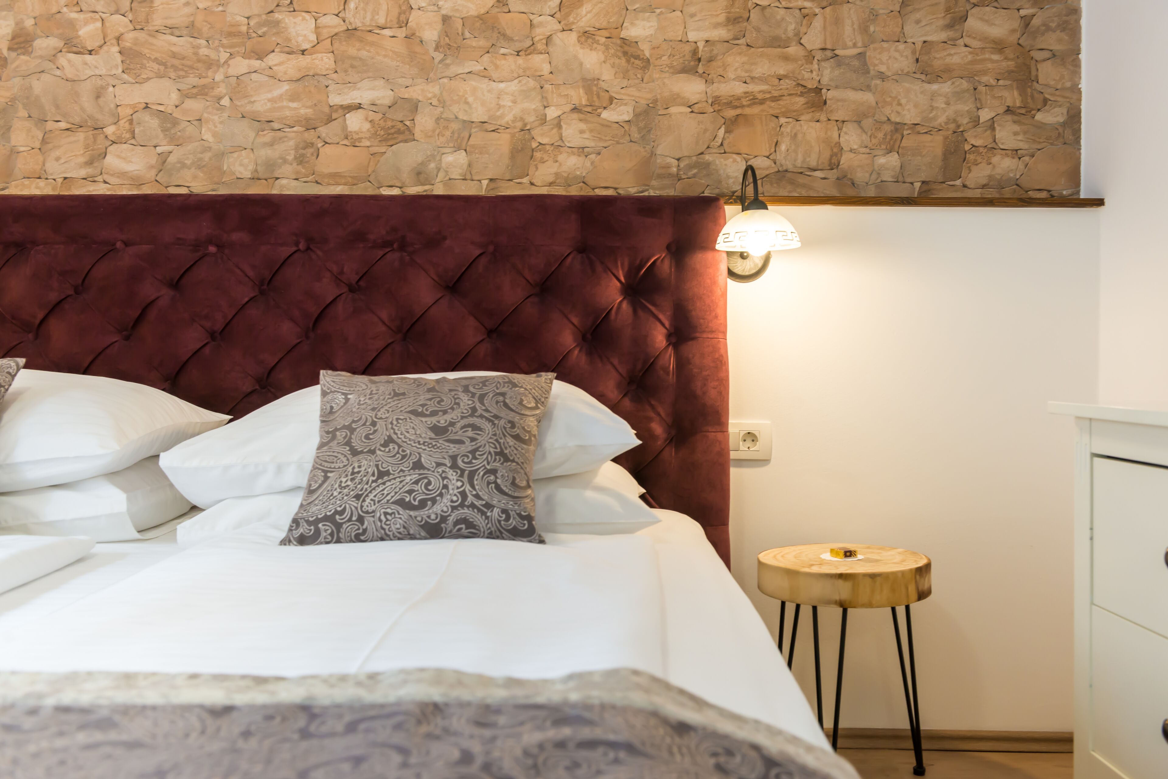 A bedroom with a stone wall, a tufted headboard, a bedside table, and a lamp.