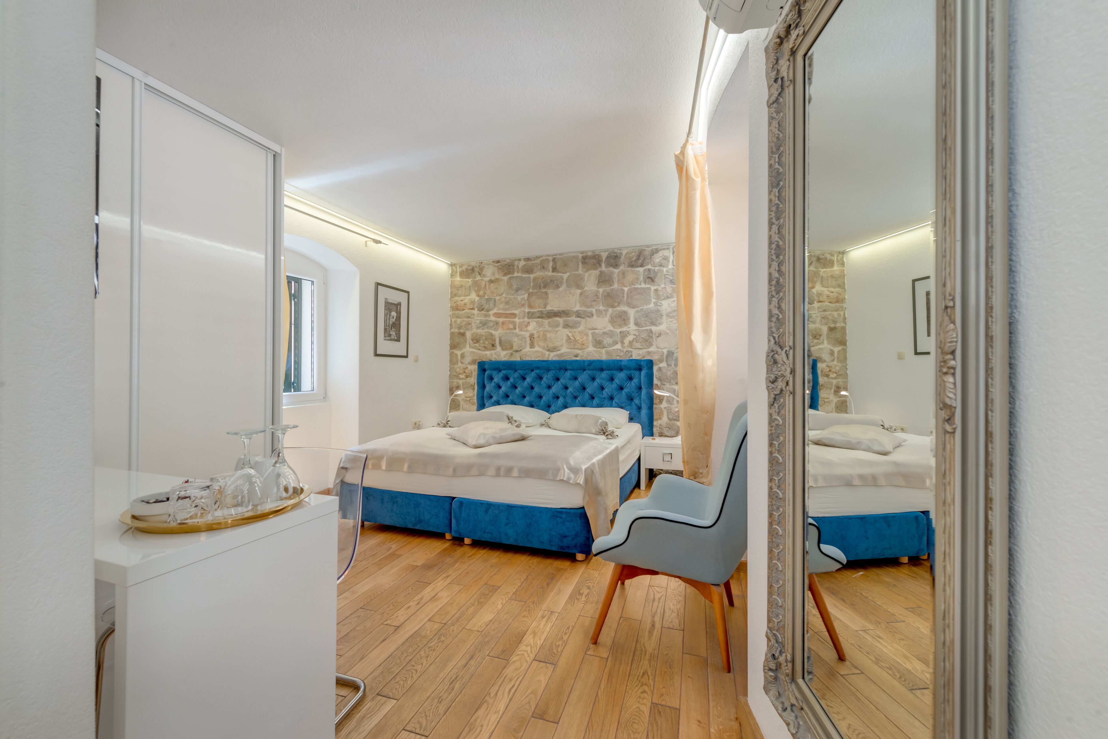 A bedroom with a stone wall, a blue upholstered headboard, a wooden floor, a white bedside table, and a chair.