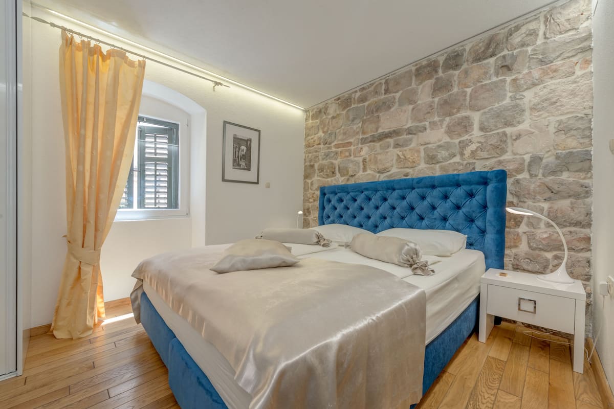 A bedroom with a stone wall, a blue headboard, a wooden floor, and a window with blinds.