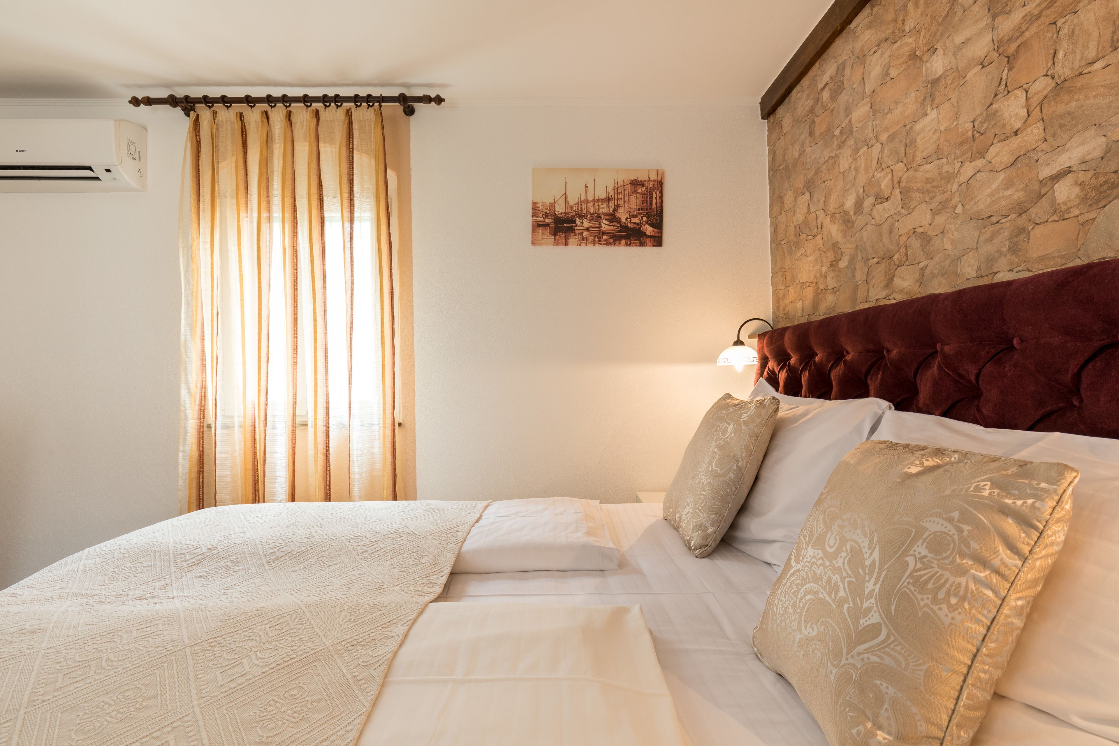A bedroom with a stone wall, a bed with white linens, a beige throw blanket, and a painting of boats.