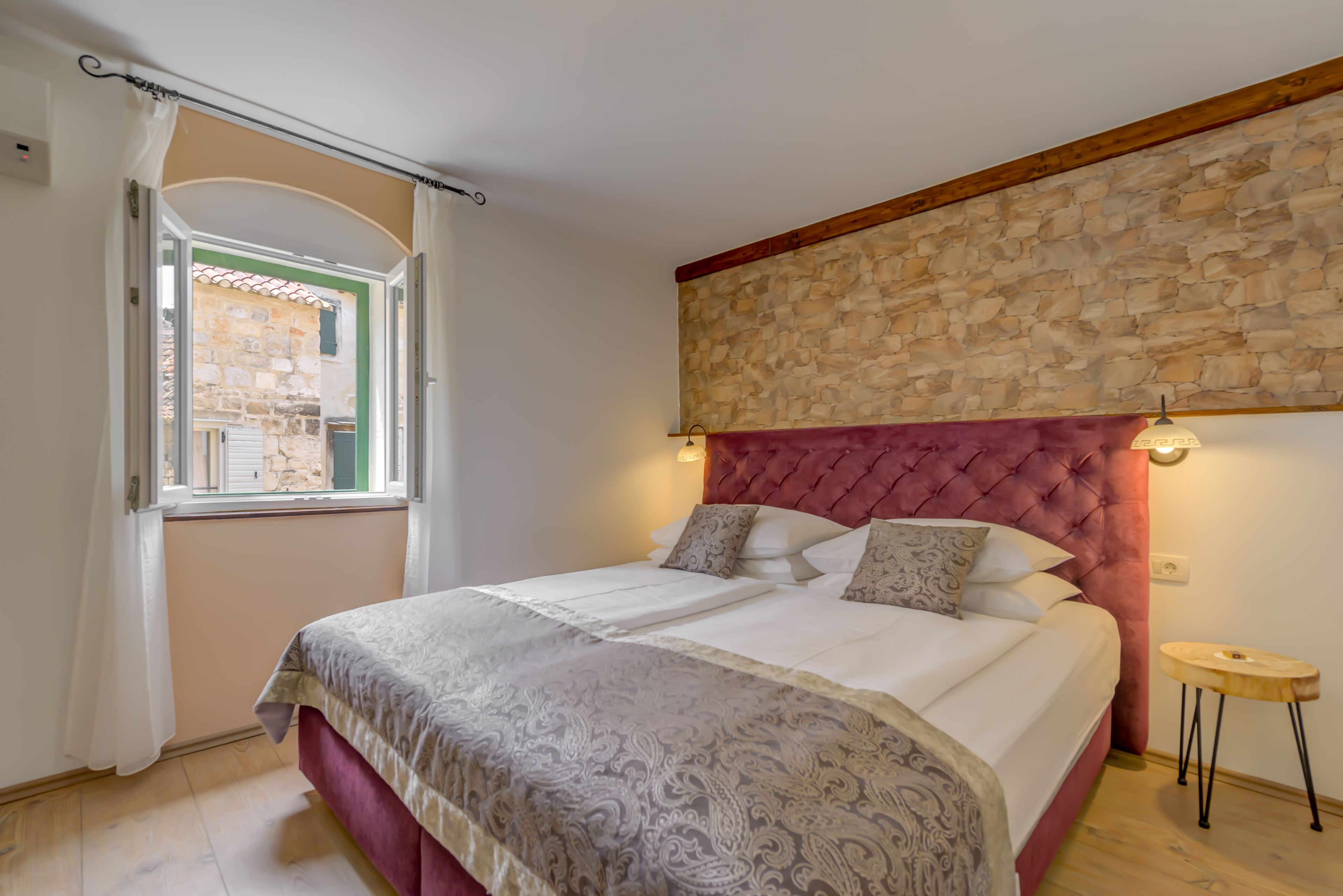 A bedroom with a stone wall, a red headboard, and a window with a view of a building.