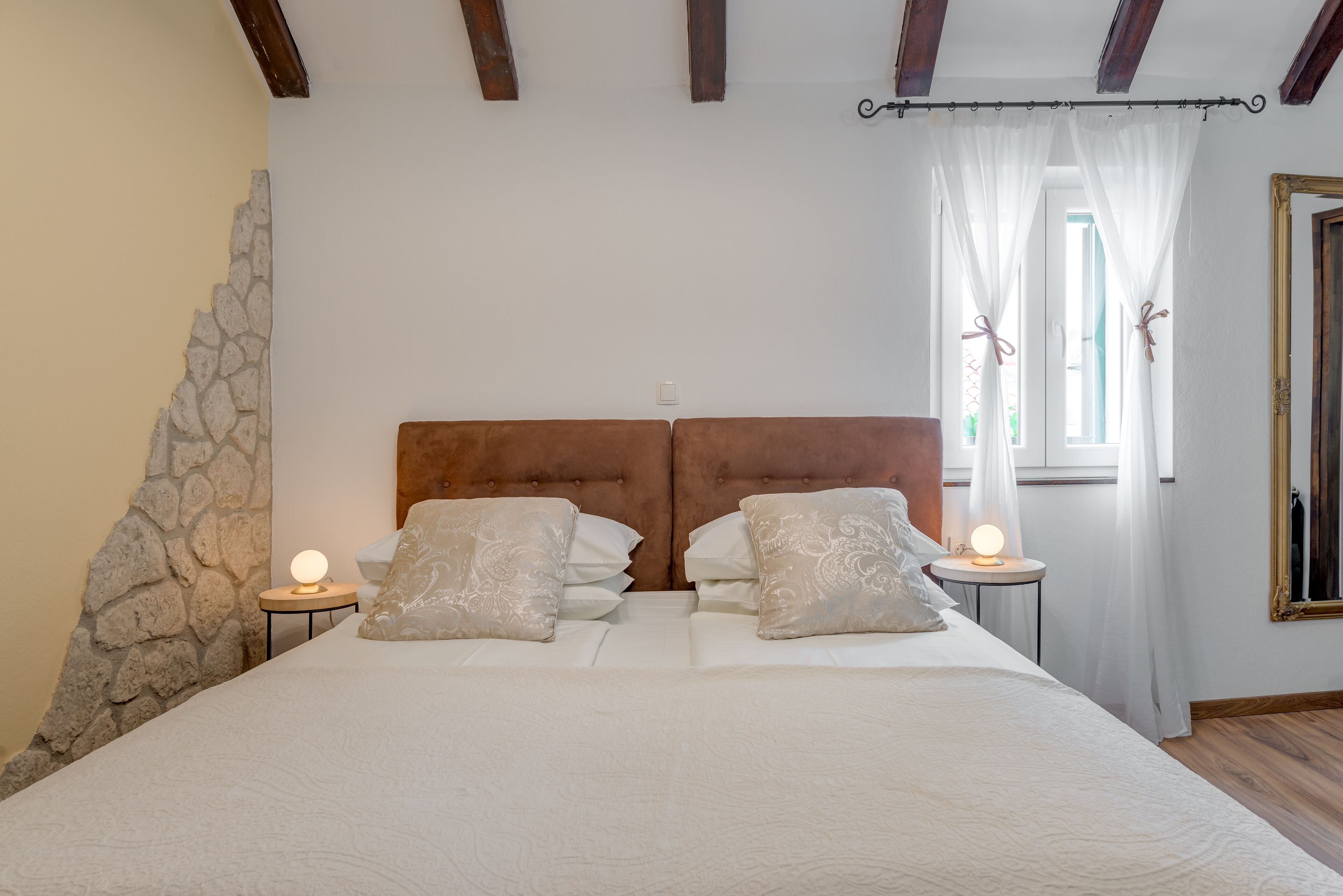 A bedroom with a stone wall, a wooden headboard, two bedside tables with lamps, and a window with curtains.
