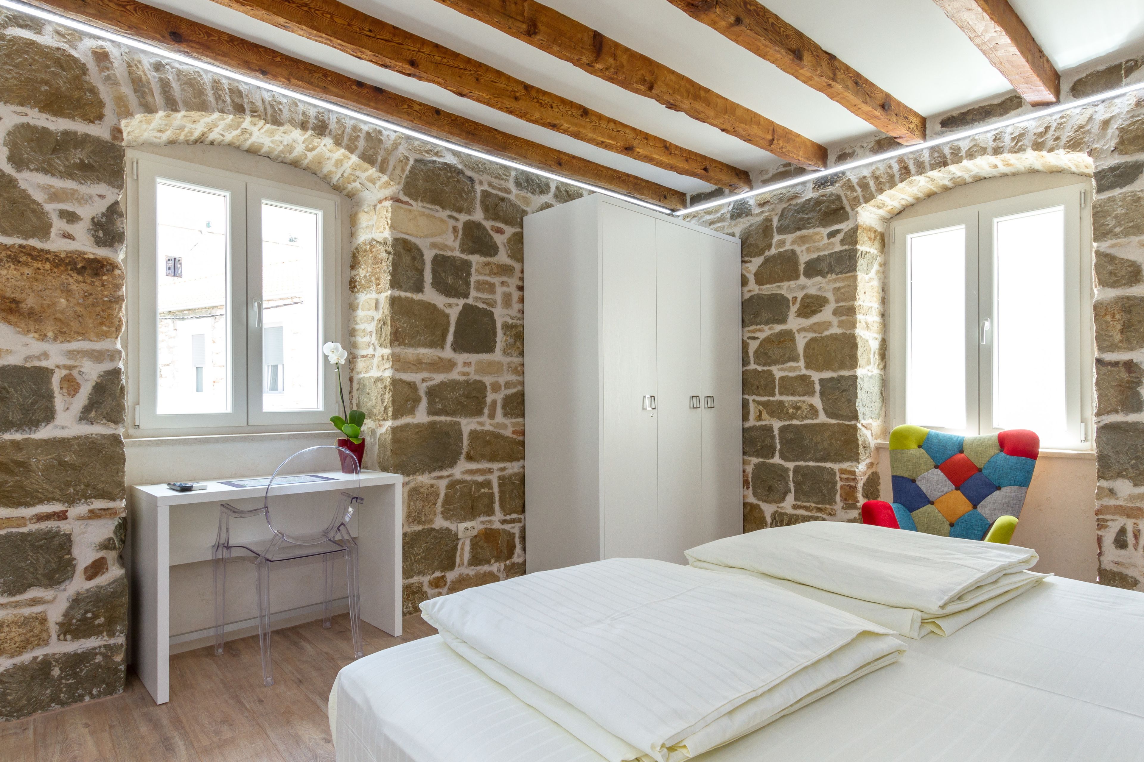 A bedroom with stone walls, a wooden ceiling, a white wardrobe, a bed with white bedding, a desk with a chair, and a colorful chair.