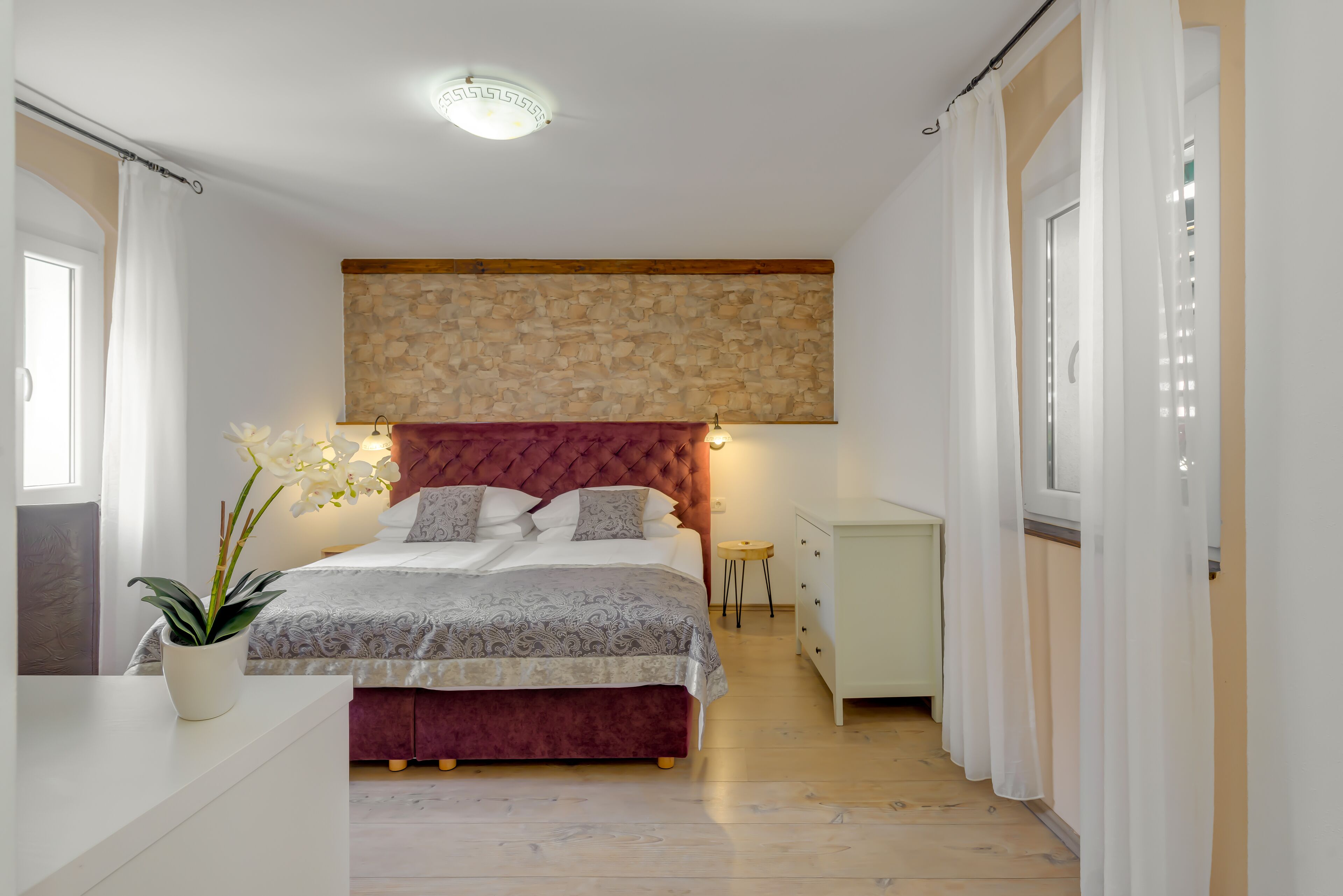 A bedroom with a large bed, a stone wall, a small bedside table, and a potted plant.