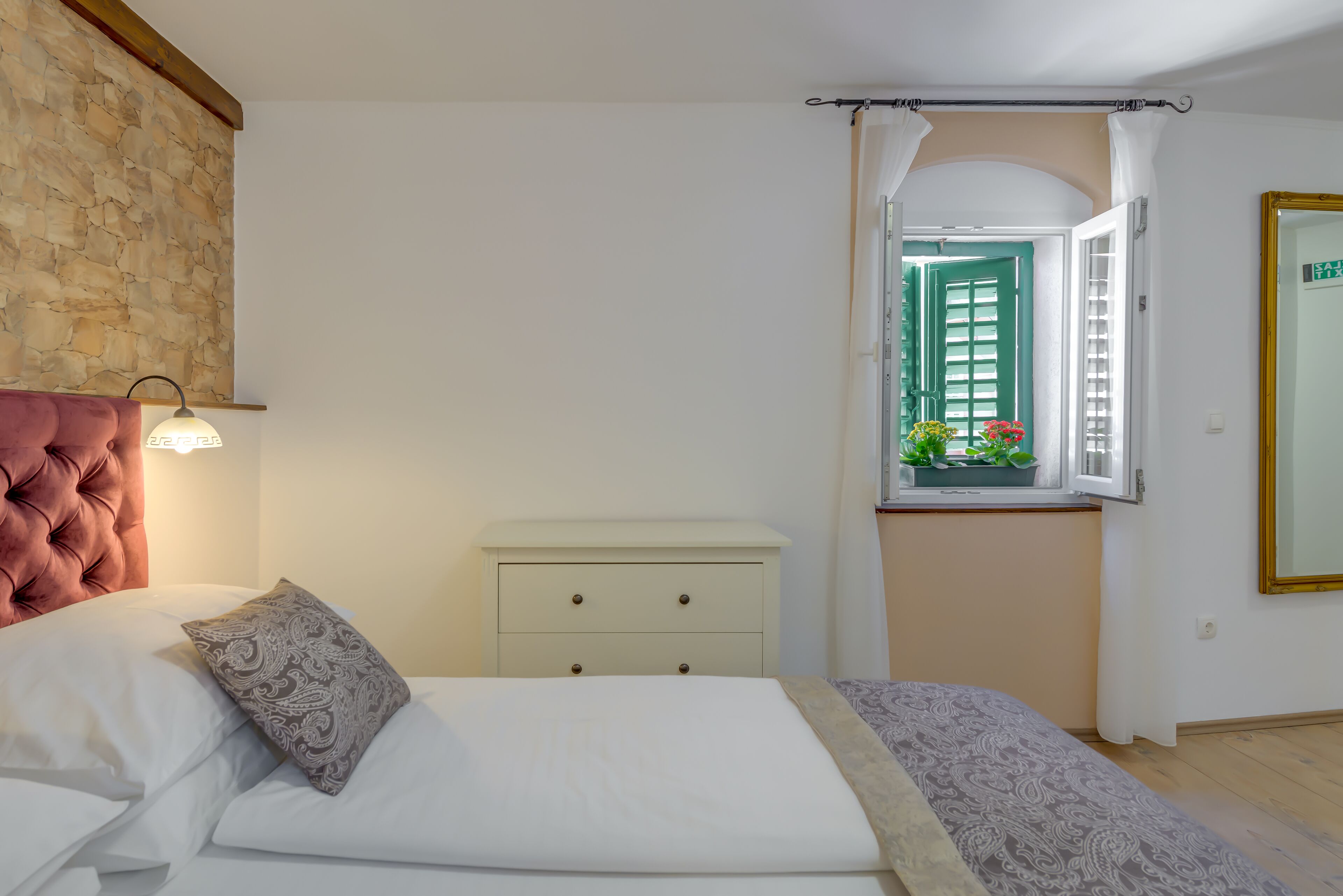 A bedroom with a bed, a nightstand, a dresser, a window with a view of greenery, and a stone wall.