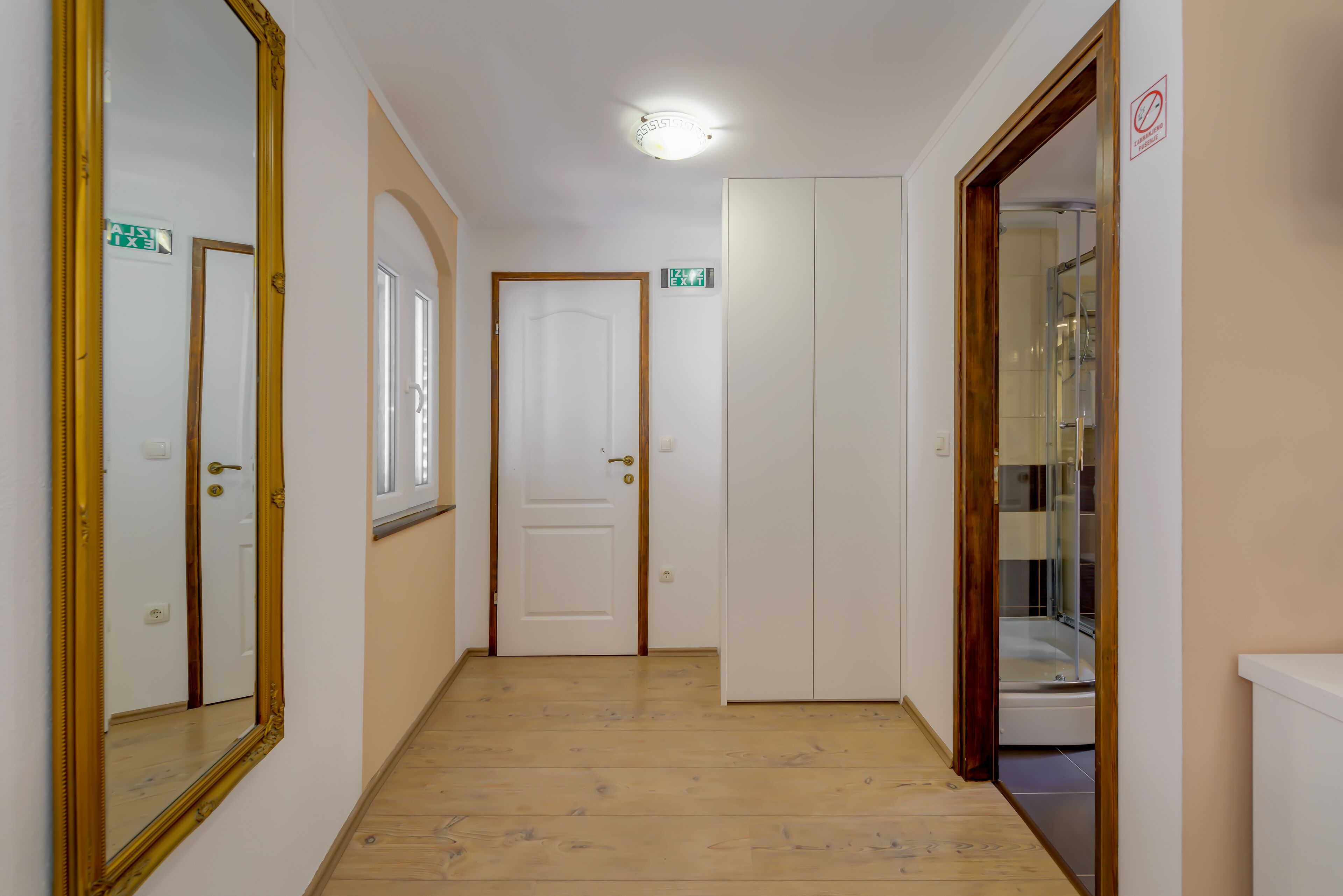 Un couloir avec un plancher en bois, des murs blancs et un grand miroir sur le côté gauche. On y trouve des portes menant à différentes pièces et un espace douche.