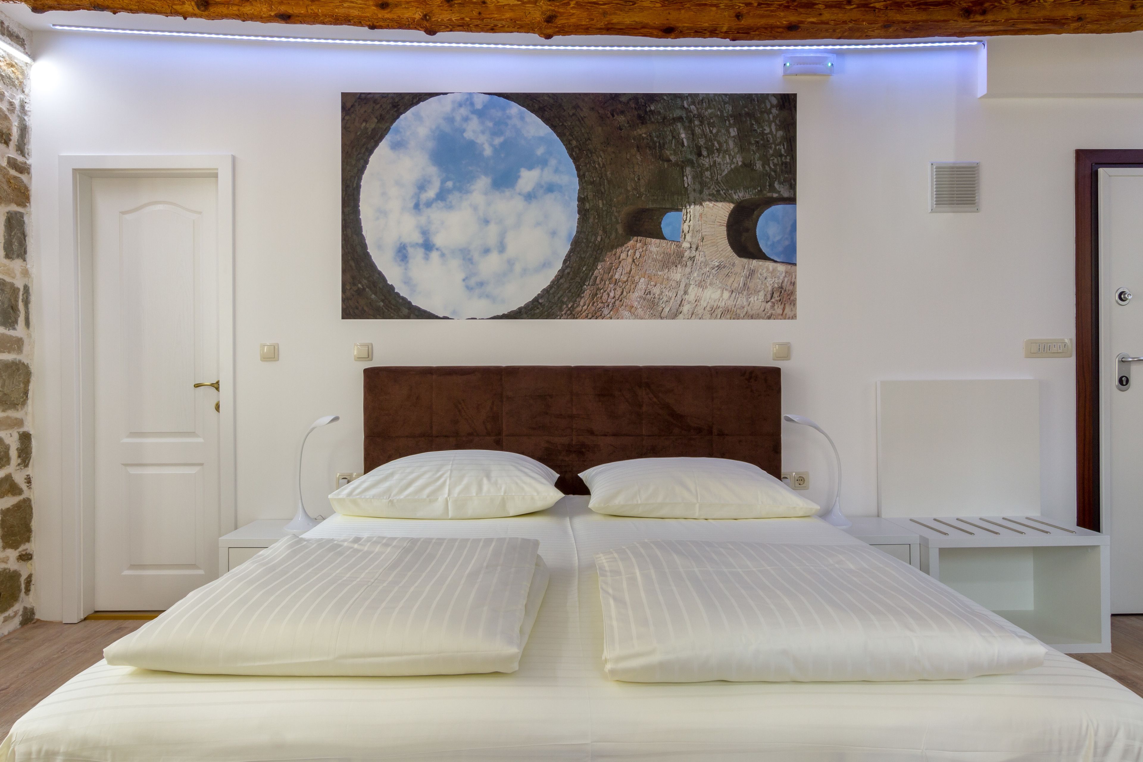 A bedroom with a large bed, two bedside tables, a nightstand, a wall-mounted lamp, and a painting of a moon and landscape.