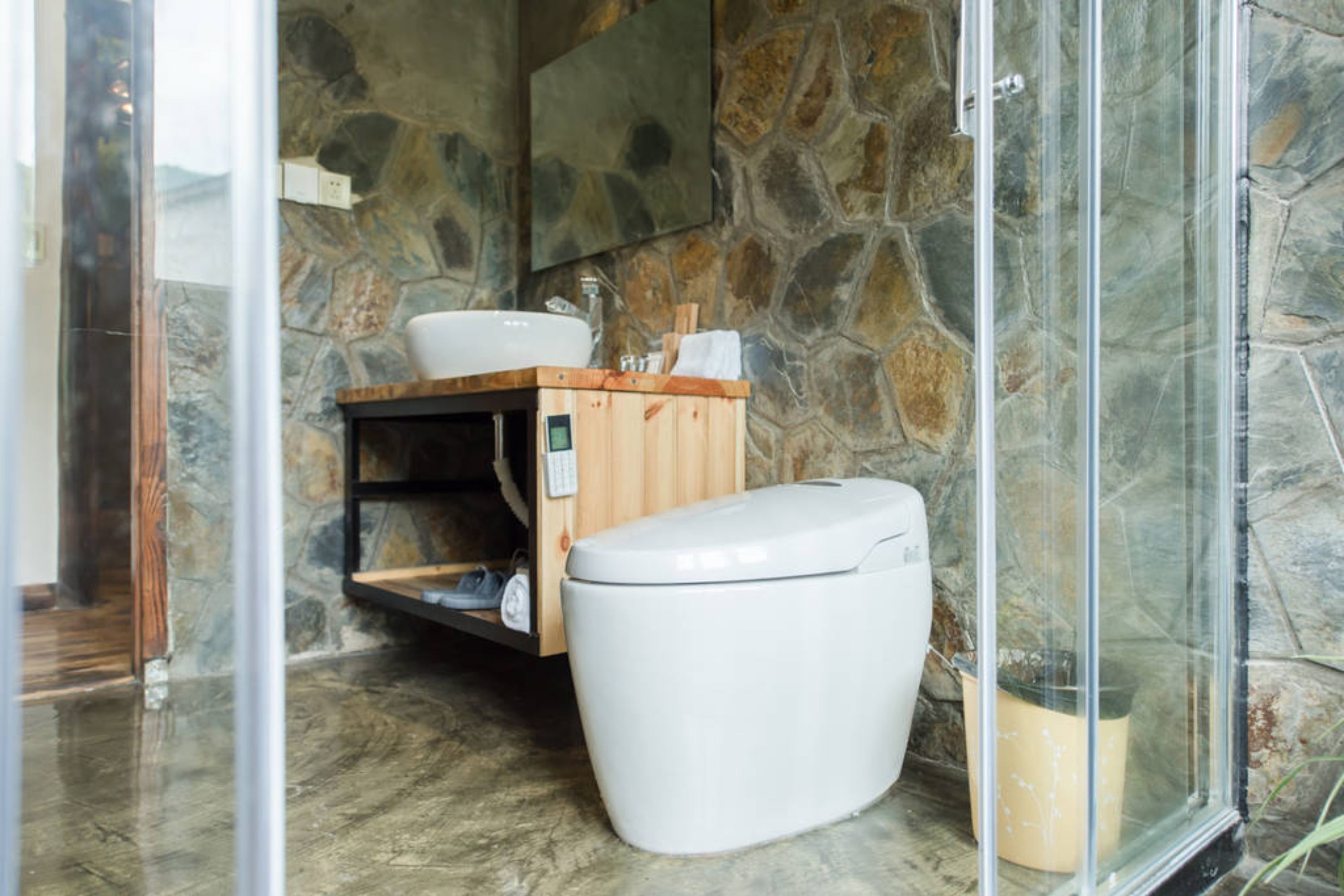 Un baño con pared de piedra, tocador de madera, inodoro blanco y cabina de ducha de vidrio.