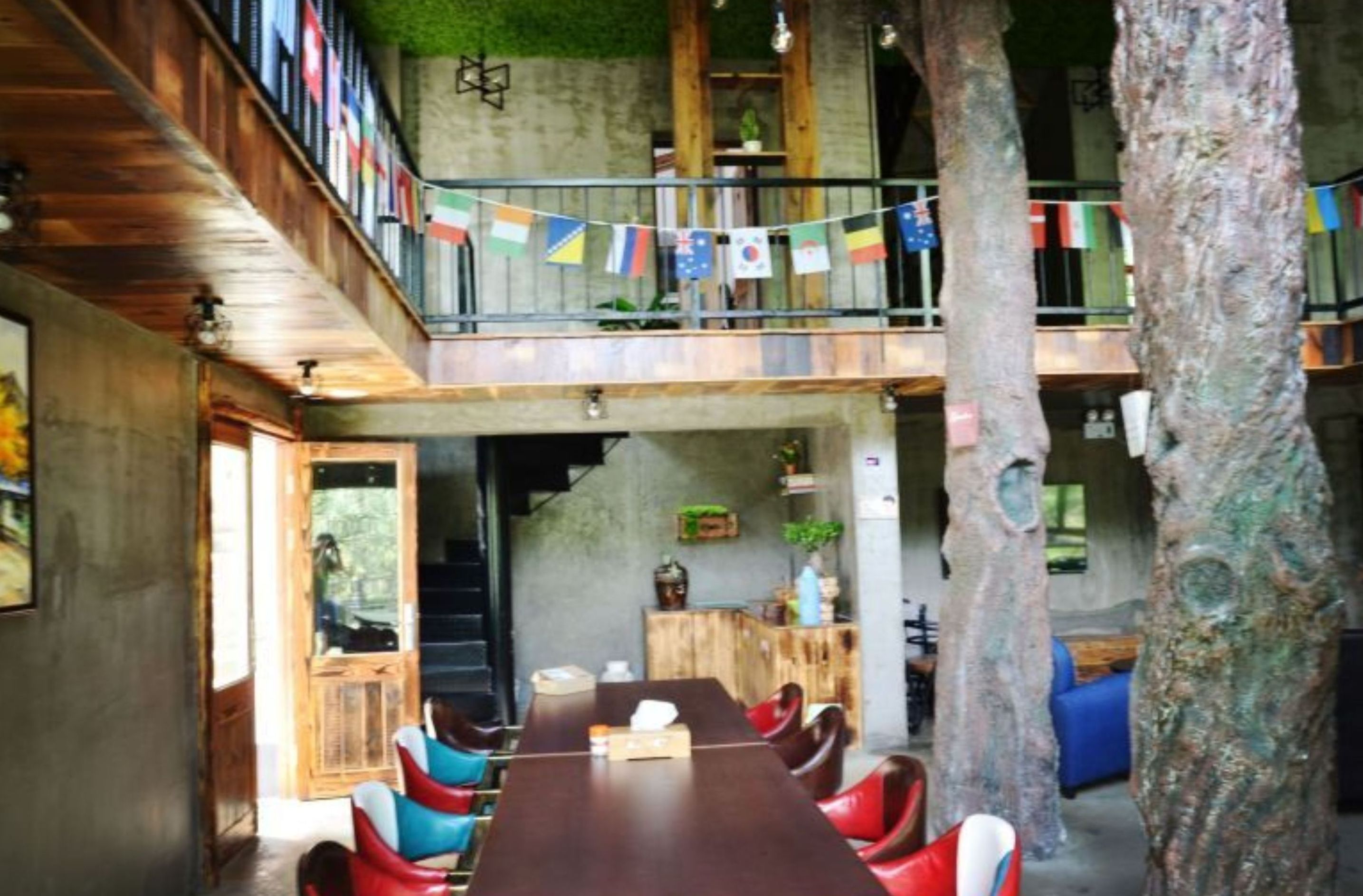 Un comedor con una mesa larga, sillas rojas y azules, y un tronco de árbol grande en el centro. Hay banderas colgadas del techo.