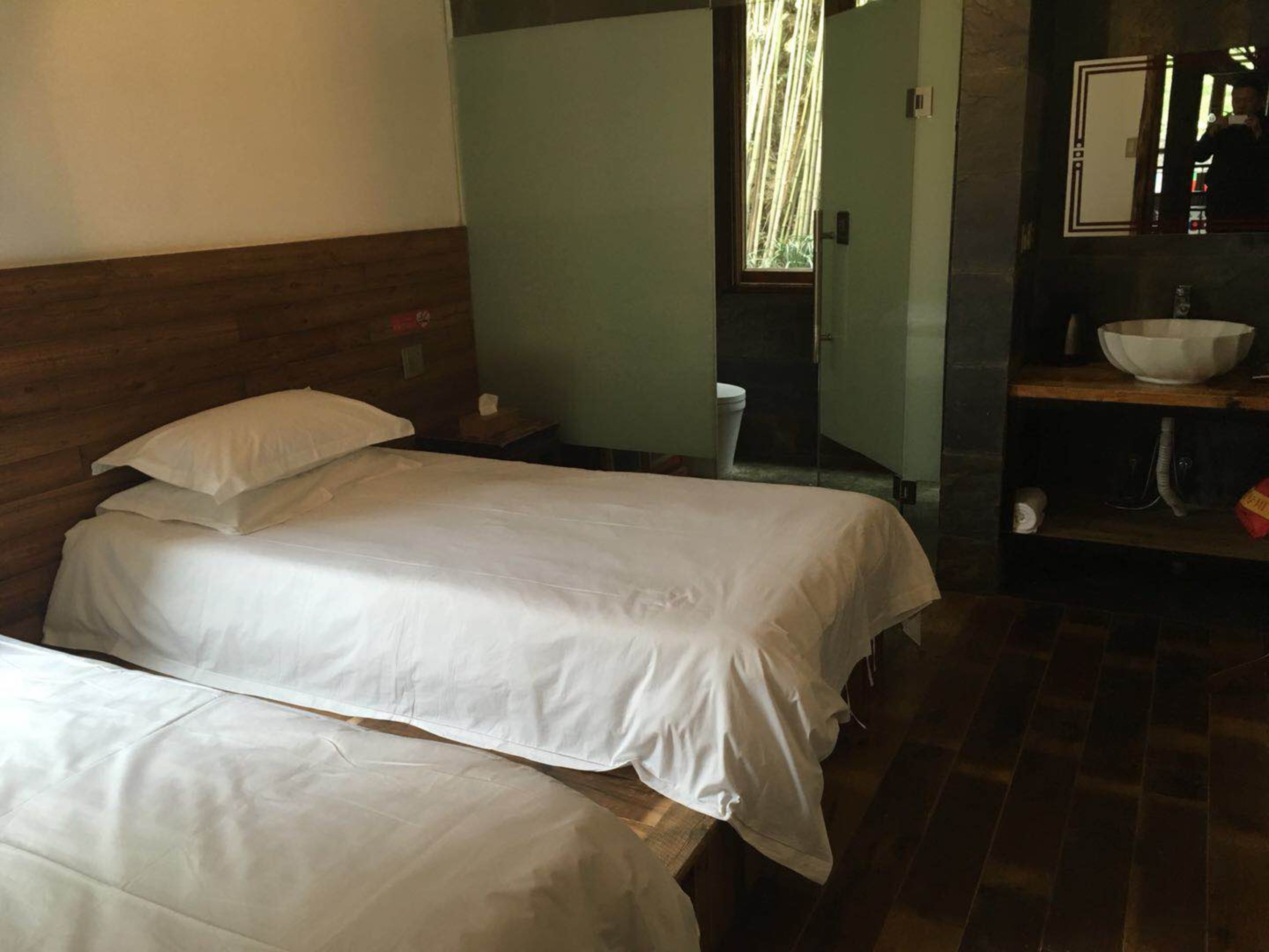Quarto com duas camas, cabeceira de madeira, pequena janela com bambu visível e pia com espelho acima.