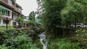 A traditional building with a thatched roof, a small stream, and lush greenery. - Moganshan Jianqi Inn (Huzhou)