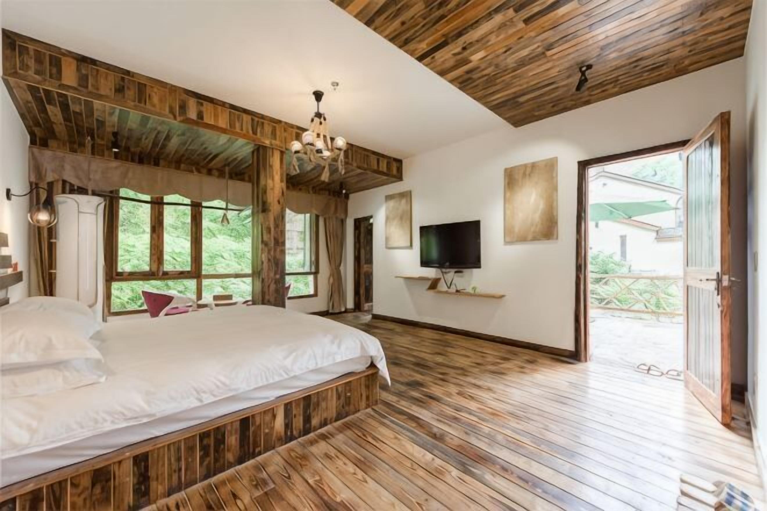 A bedroom with a large bed, wooden flooring, a TV, and a view of greenery.