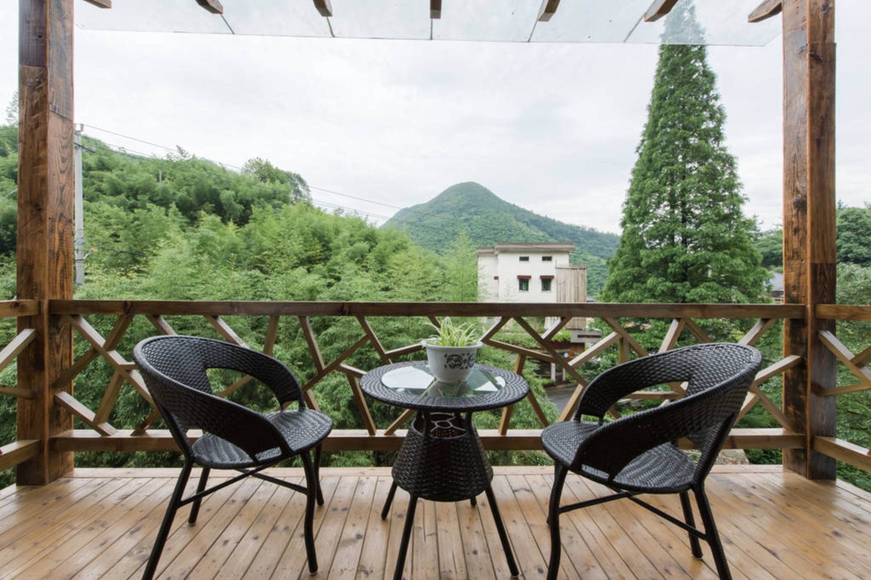 A wooden deck with two wicker chairs and a glass table, overlooking a mountainous landscape with a building in the distance.