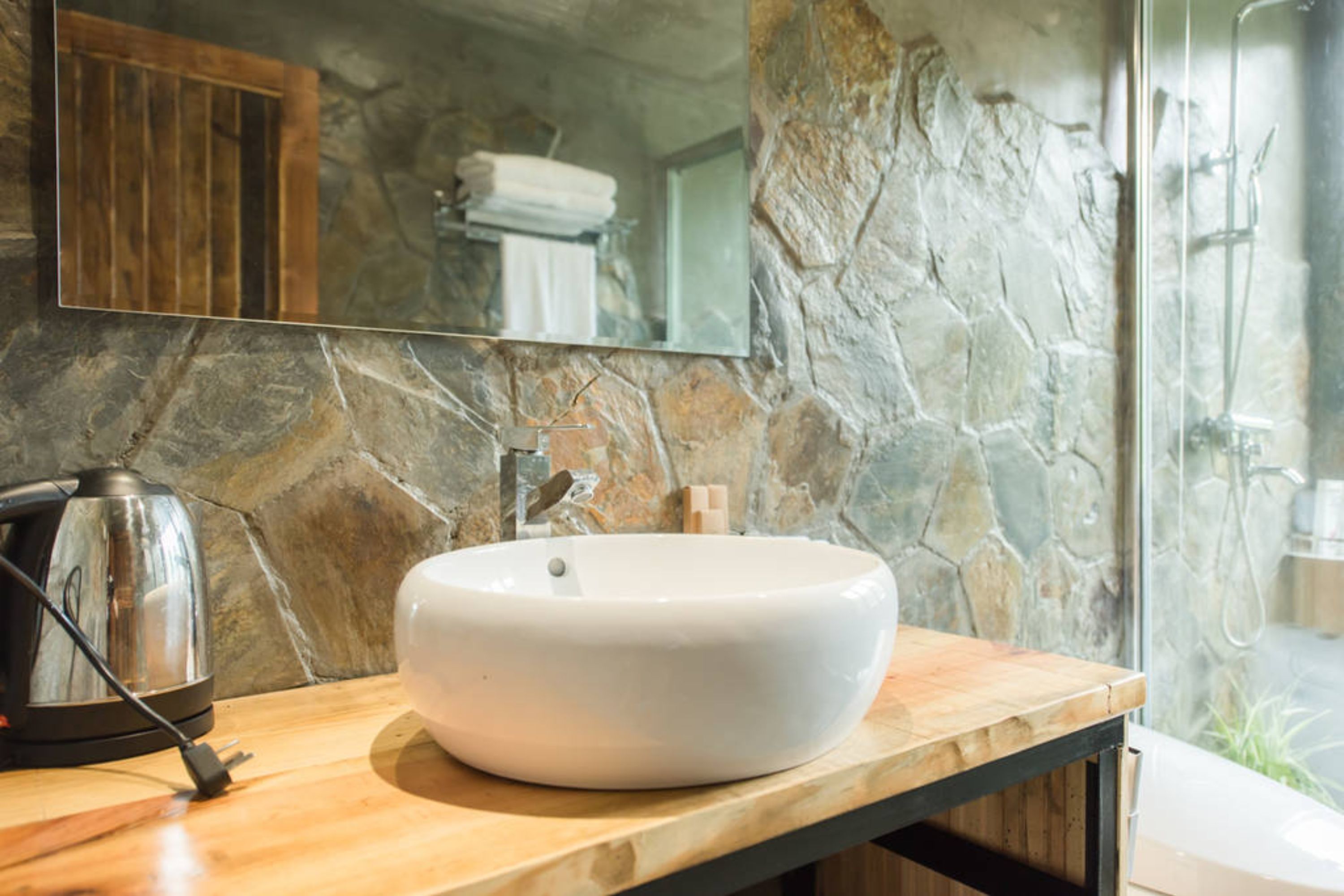 A bathroom with a stone wall, a wooden countertop, a white sink, a mirror, and a shower area.