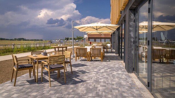 Een terras buiten met houten tafels en stoelen, parasols en uitzicht op de lucht en het landschap.