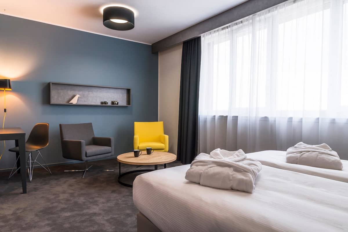Une chambre d’hôtel comprenant un lit, deux tables de chevet, un bureau, une chaise, une petite table ronde et une étagère murale agrémentée d’objets décoratifs.
