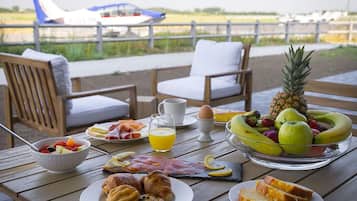 Una mesa de desayuno con croissants, fruta, huevos y jugo, con vista a una playa y un avión al fondo.