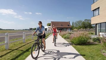 Duas pessoas andando de bicicleta numa calçada, com um edifício e uma cerca ao fundo.