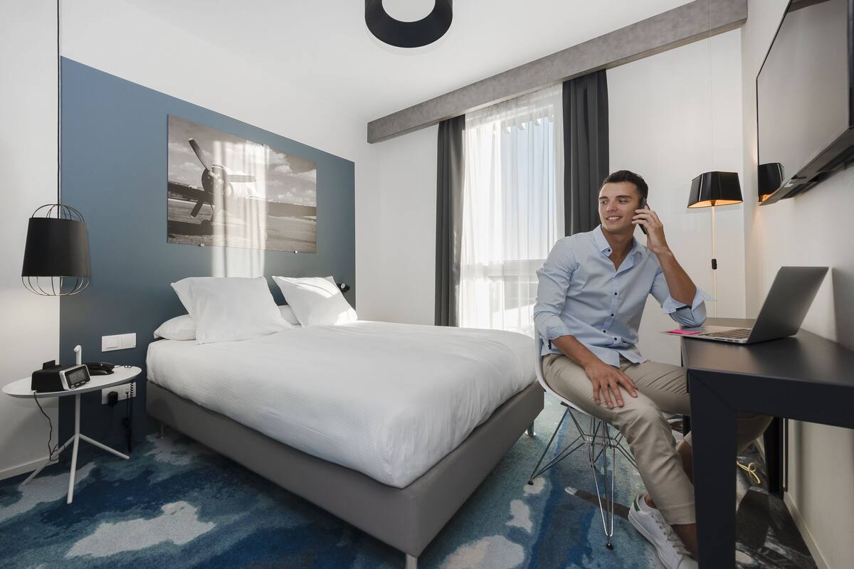 Une chambre d’hôtel moderne équipée d’un lit, d’un bureau avec un ordinateur portable, d’une table de chevet avec un téléphone et d’une grande fenêtre avec des rideaux.