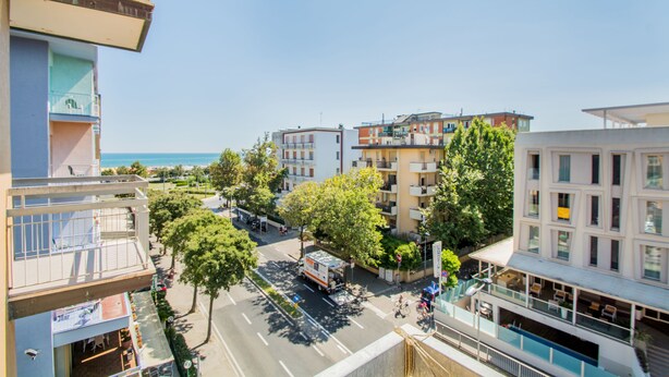 A multi-story building with balconies, a street with cars and bicycles, and a clear view of the sea in the background.