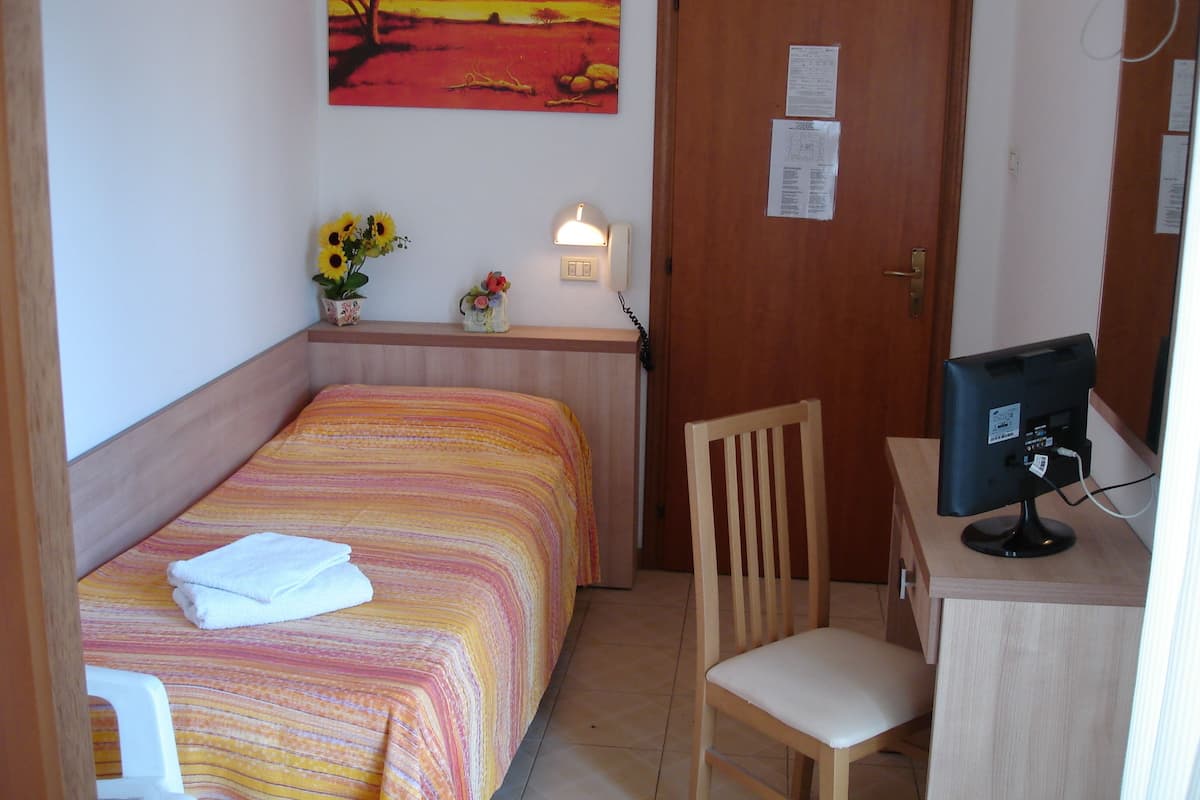 A small room with a bed, a desk with a computer, a chair, a wall-mounted phone, and a painting of a sunset.