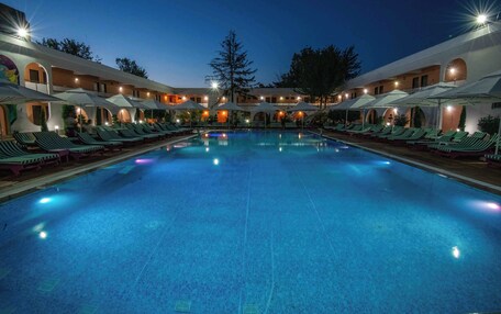 A large indoor swimming pool with lounge chairs and umbrellas, surrounded by a building with multiple balconies and a central tree.