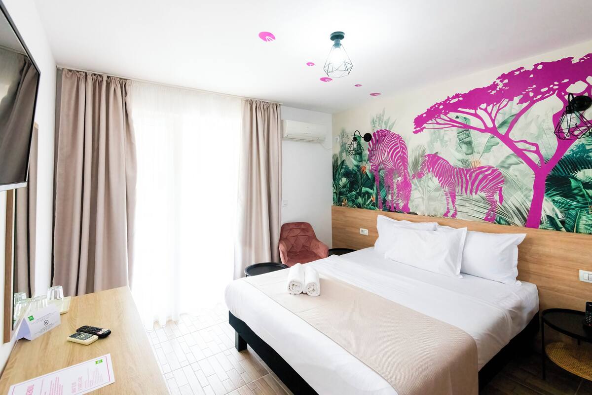 A hotel room with a bed, a desk with a phone and a menu, a chair, and a wall mural.