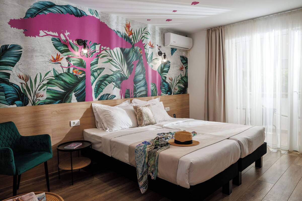 A hotel room with a bed, a green chair, a small table, and a wall mural.