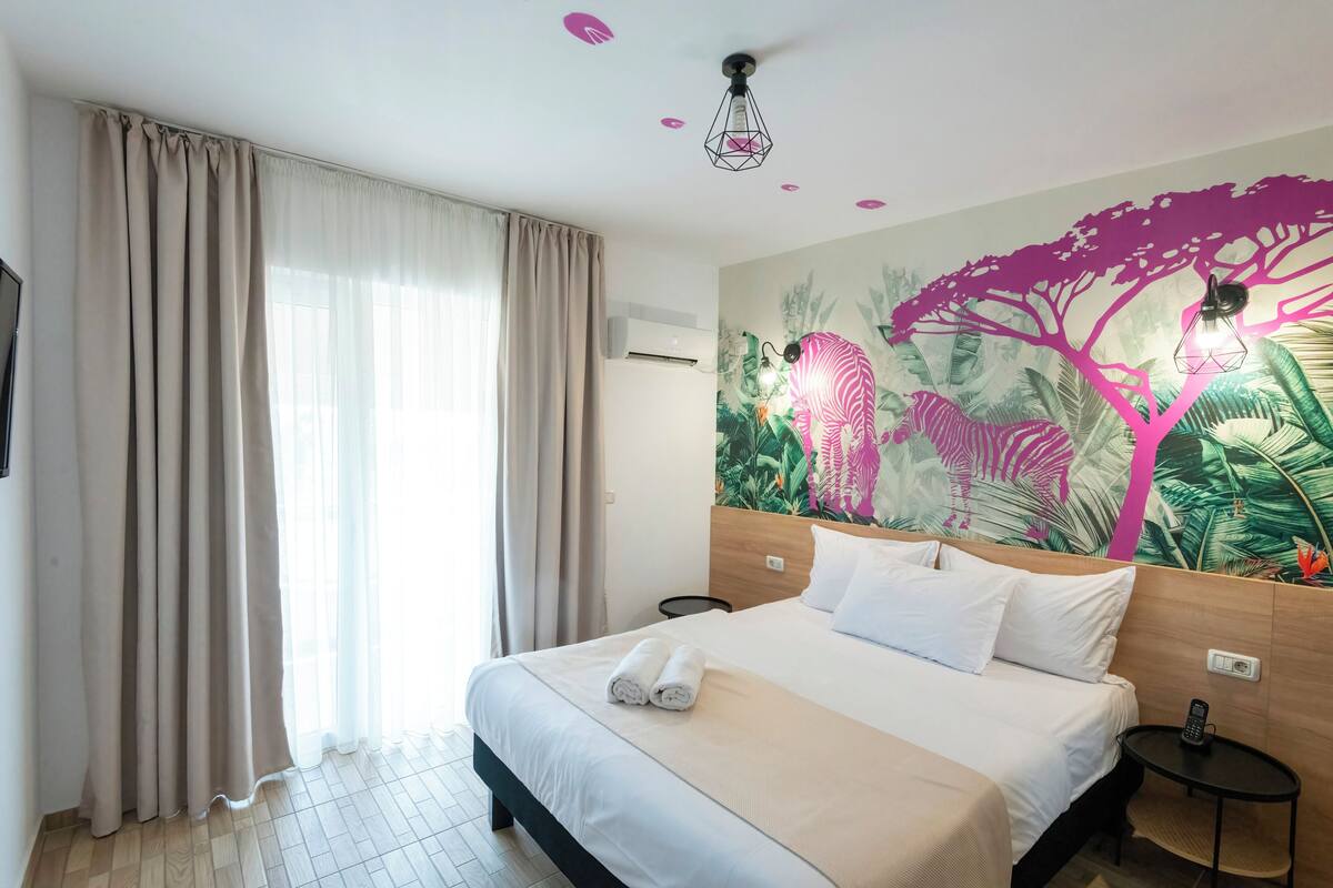 A hotel room with a bed, curtains, a wall mural, a ceiling light, and a small table with a remote control.