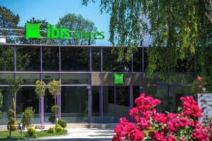A modern building with a green 'ibis styles' sign, large windows, and a well-maintained garden with small trees and flowers. - ibis Styles Venus (Venus)