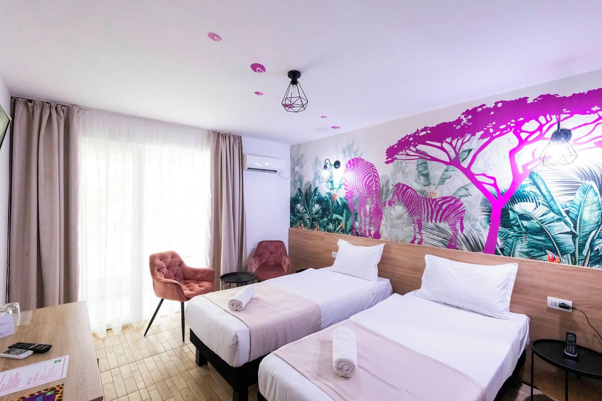 A hotel room with two beds, a desk, a chair, and a mural of a tree and animals on the wall.