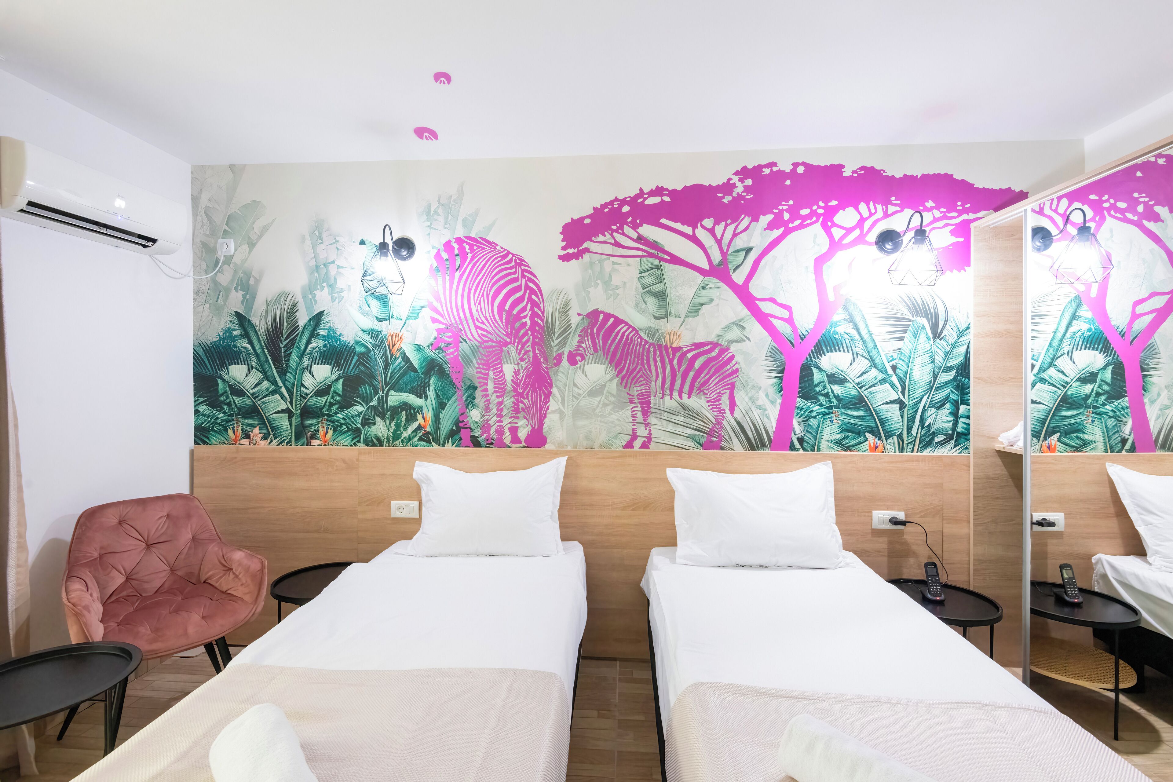 A hotel room with two beds, a pink chair, a small table, and a wall mural with a zebra and trees.