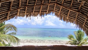 A beach view from a thatched-roof structure with clear turquoise water and palm trees. - Zanziblue (Matemwe)