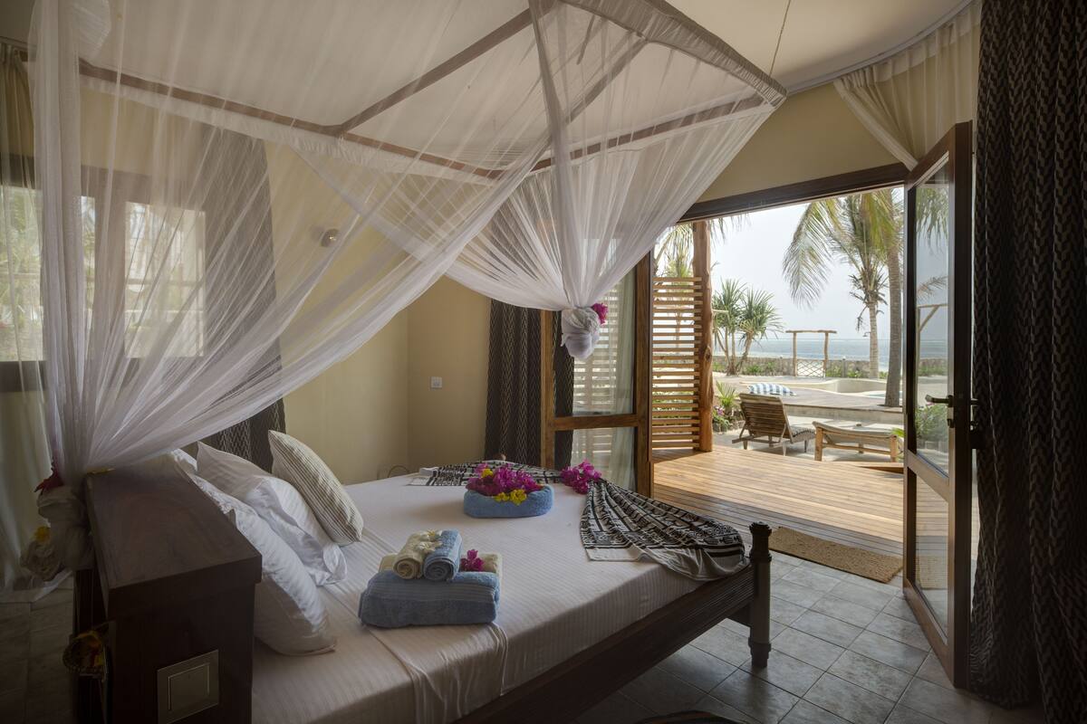 A bedroom with a bed, a bedside table, a window with a view of the beach, and a balcony with outdoor furniture.