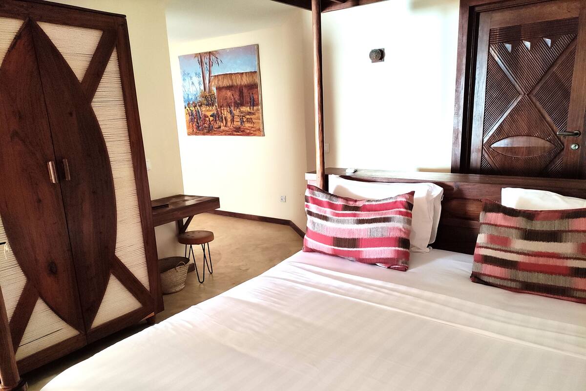 A bedroom with a bed, pillows, a wooden wardrobe, a desk, and a painting on the wall.
