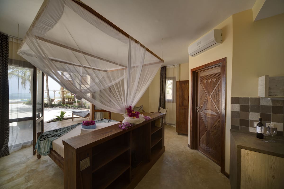A room with a bed, a bedside table, a wooden door, and a view of the beach.