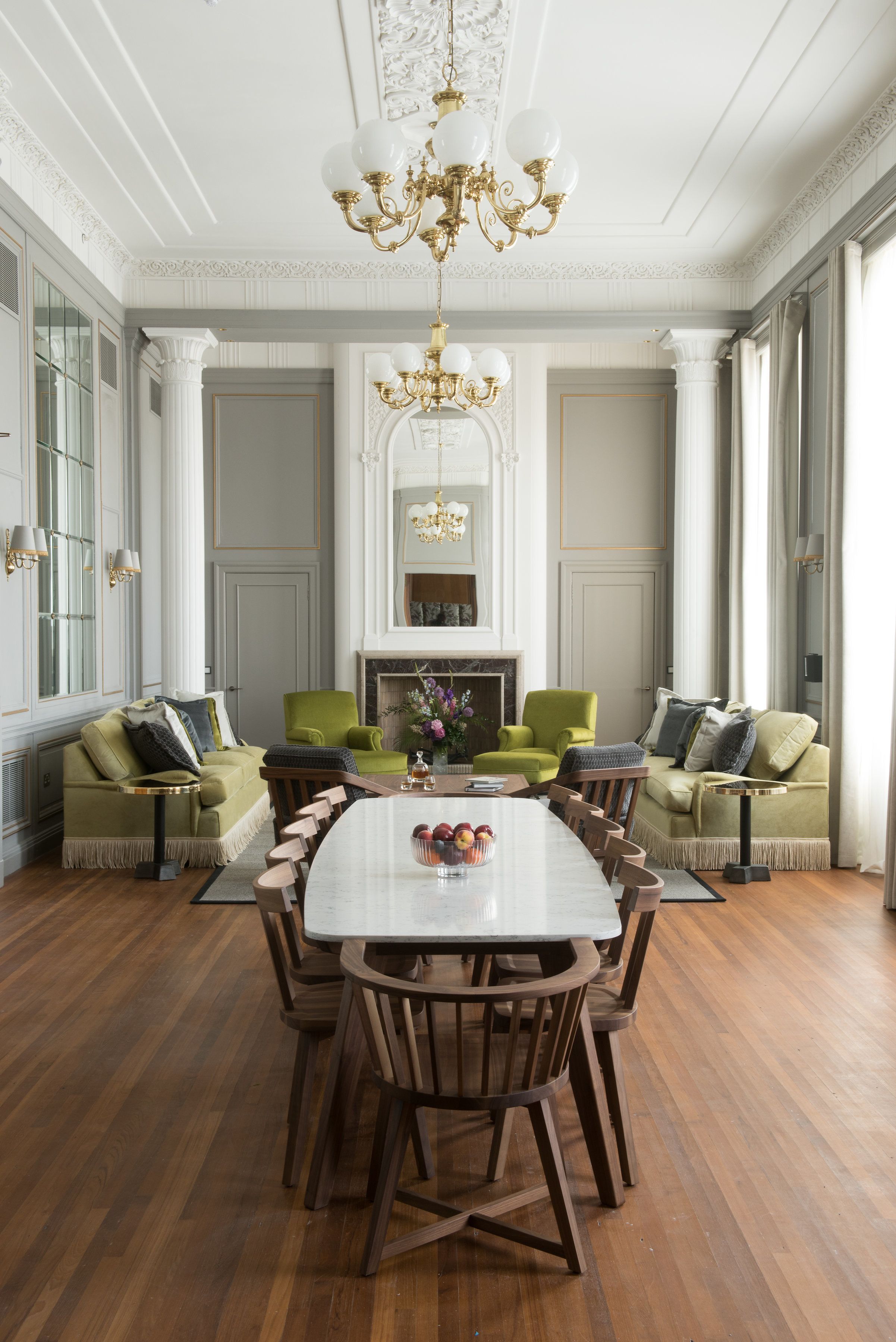 Un coin repas avec une grande table entourée de chaises en bois, flanqué de fauteuils verts et d’un canapé. Un lustre est suspendu au plafond, et une cheminée est visible au fond.