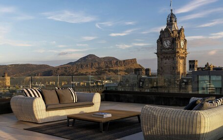 A rooftop terrace with wicker furniture, a view of a clock tower, and distant mountains.