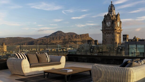 A rooftop terrace with wicker furniture, a view of a clock tower, and distant mountains. - The Edinburgh Grand, a Luxury Collection Hotel, Edinburgh (Edinburgh)