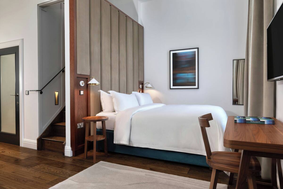 A modern hotel room with a bed, a wooden desk, a chair, a staircase, and a painting on the wall.