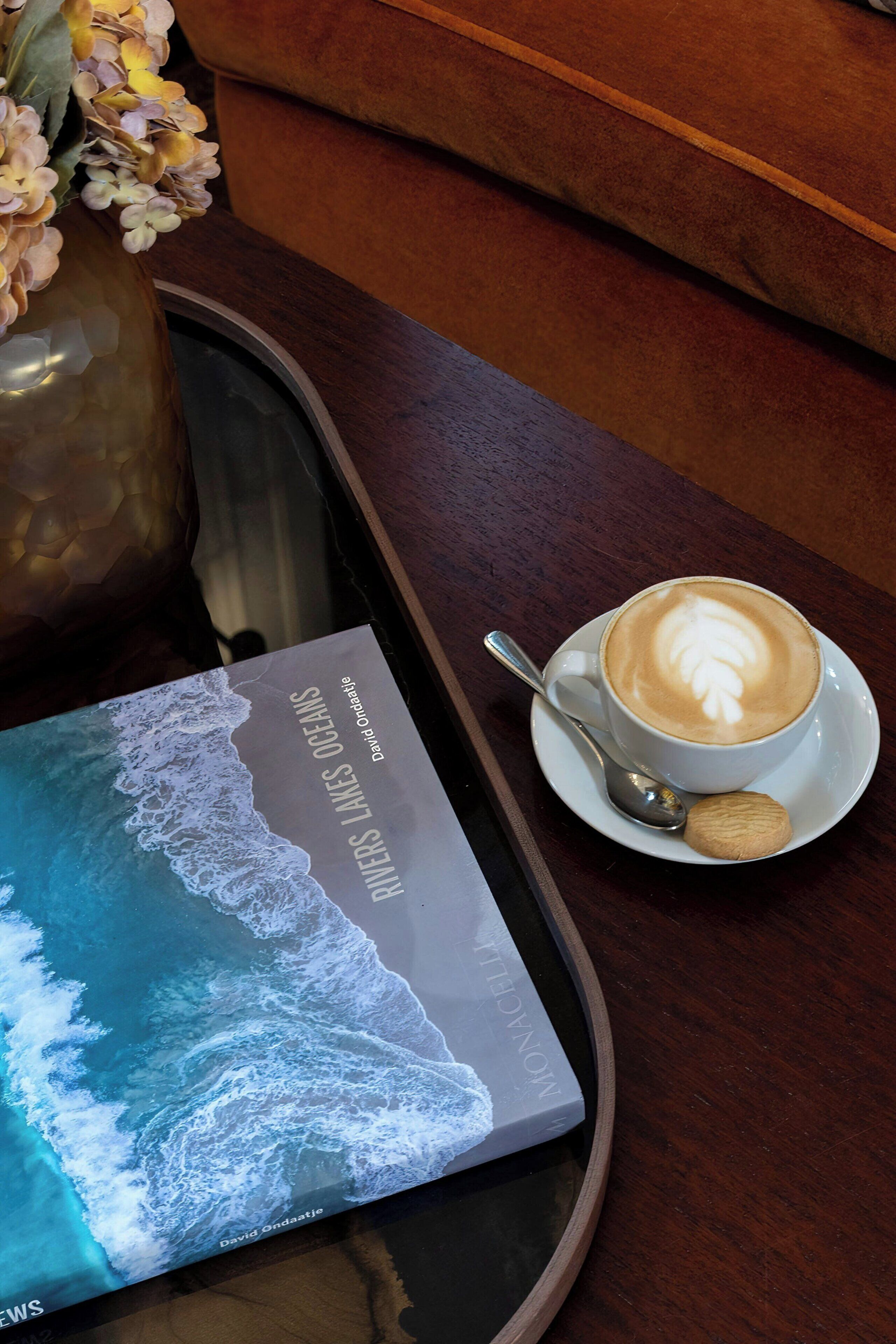 A tray with a book, a cup of coffee, and a cookie on a wooden table.