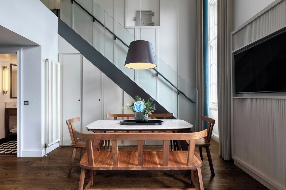 A dining area with a round table, wooden chairs, a black lamp, and a staircase in the background.