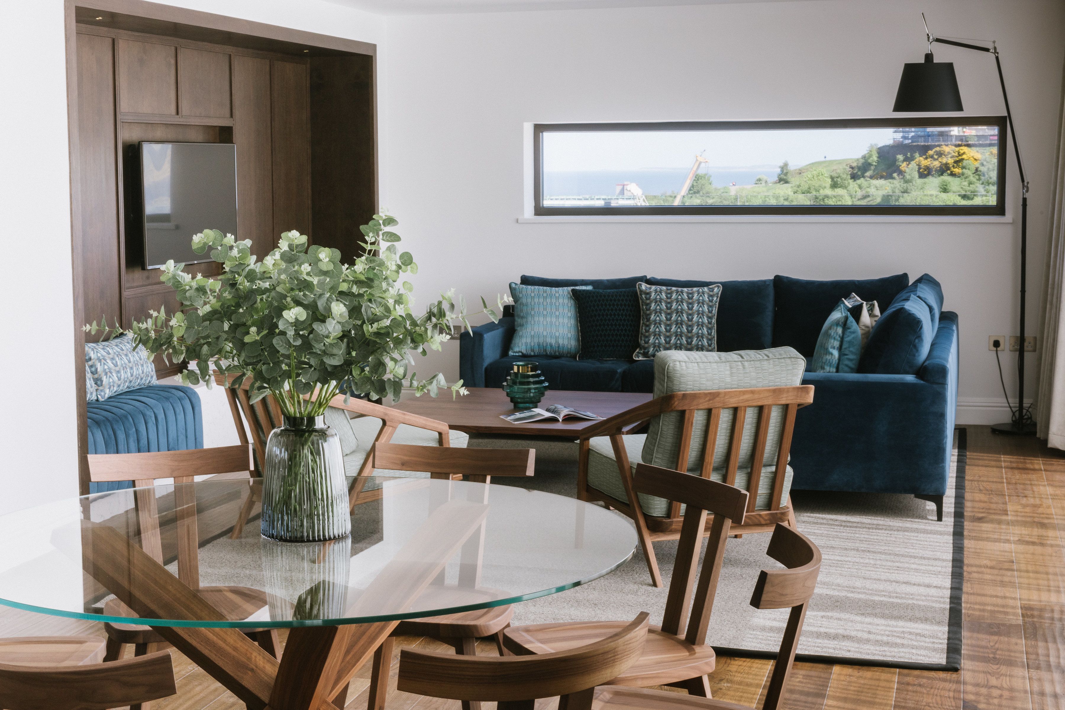 Un salón moderno con una mesa de comedor de vidrio, sillas de madera, un sofá azul con cojines estampados y vista al mar a través de un ventanal grande.