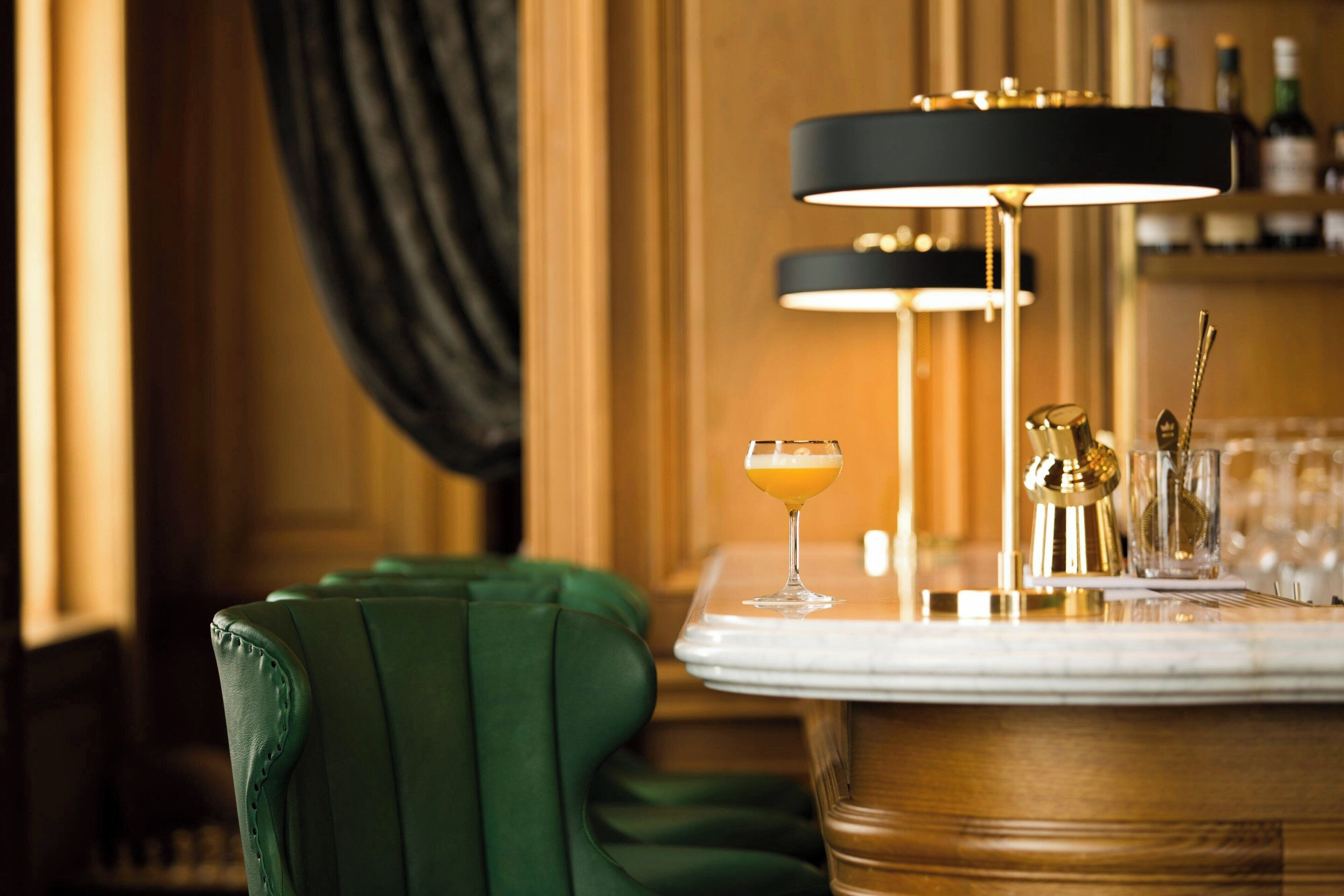 A bar area with a green upholstered chair, a marble table, and a black and gold bar lamp.
