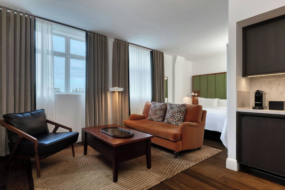 A modern hotel room with a leather armchair, a sofa with patterned cushions, a coffee machine, and a view of the cityscape through large windows.