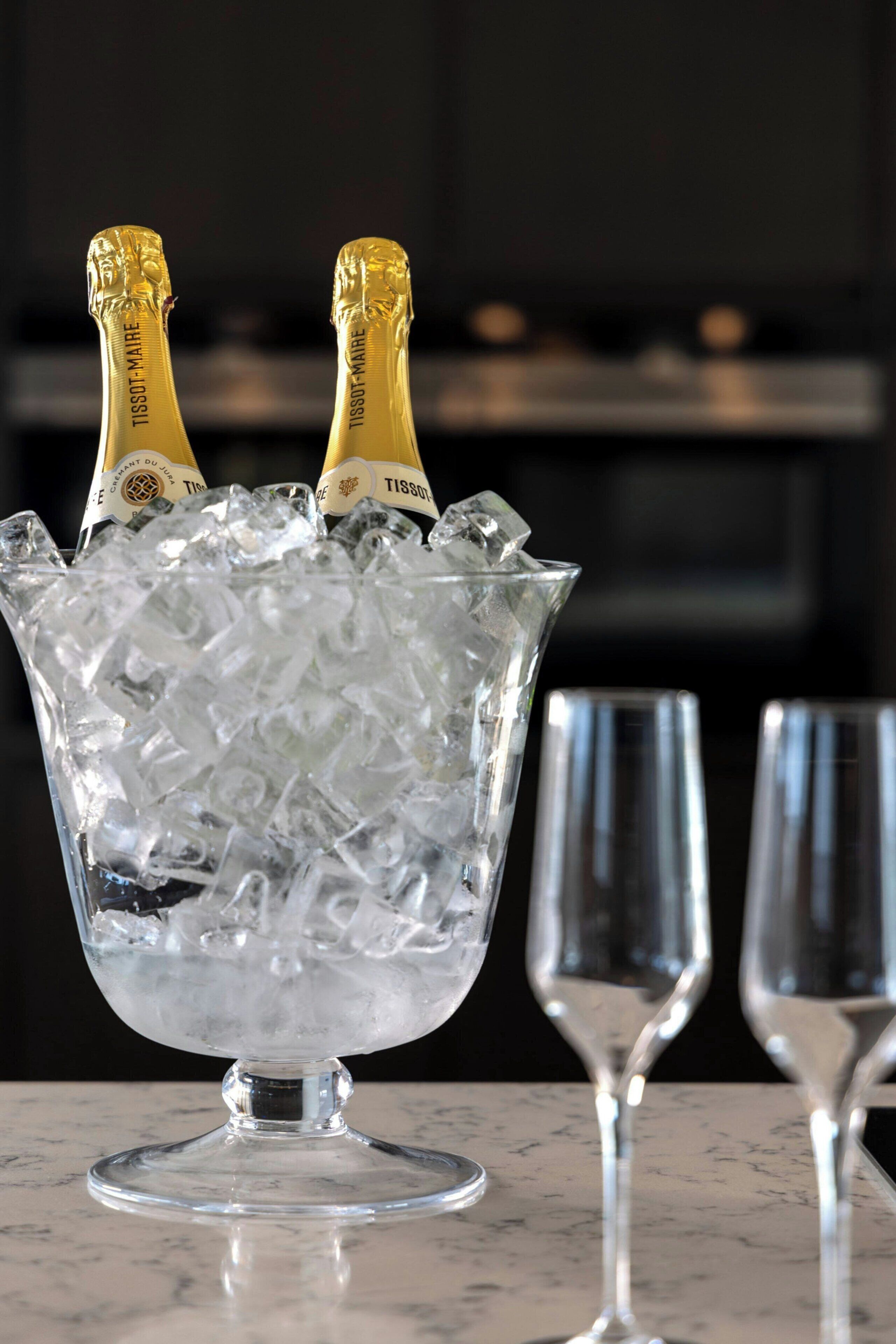 Eine Glaskugel mit Eiswürfeln und zwei mit 'TISSOT-MAIRE' beschrifteten Champagnerflaschen auf einer Marmorplatte.