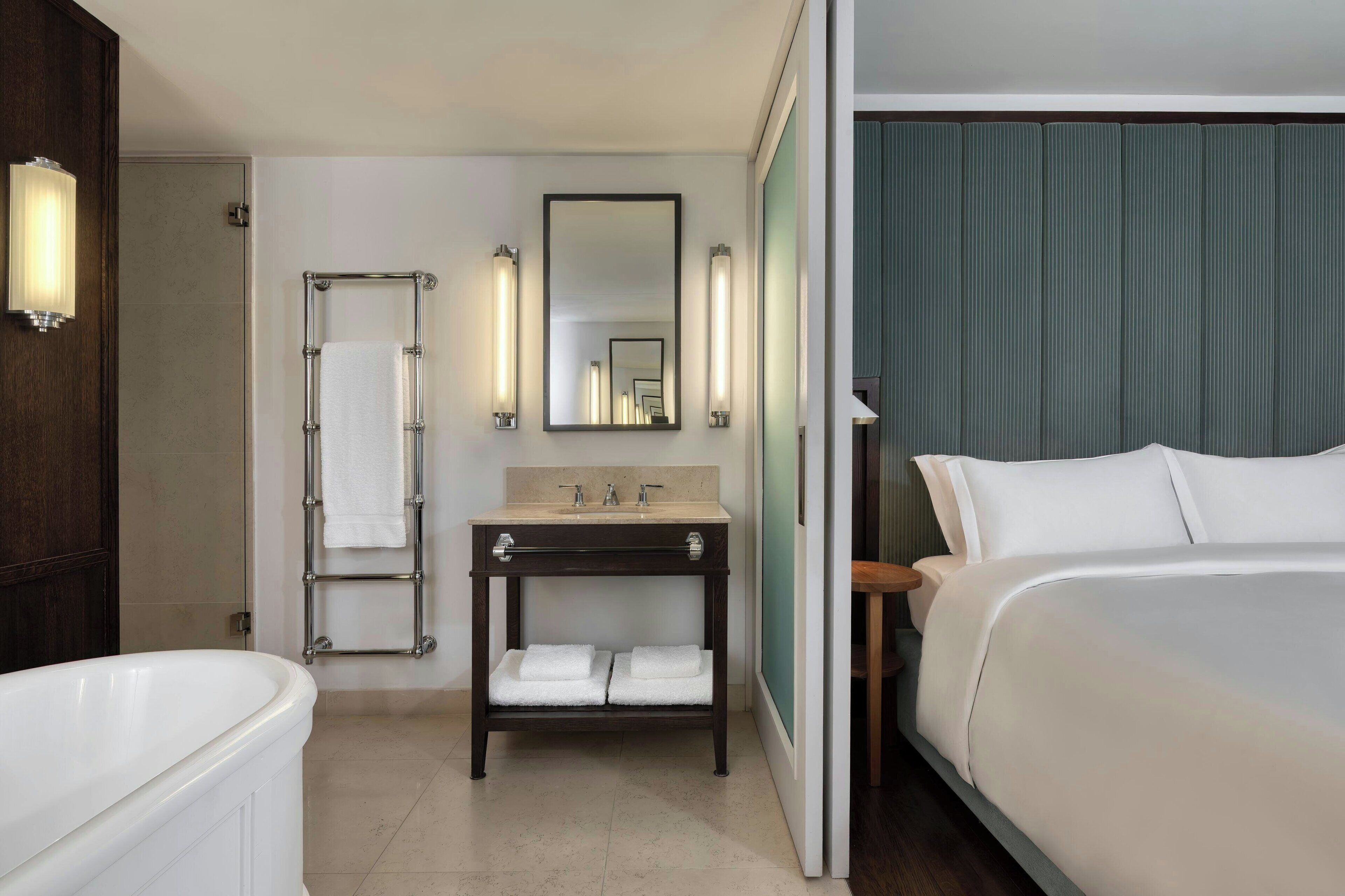 Une chambre d’hôtel avec une baignoire, une coiffeuse avec un miroir et des lampes, un lit et une vue sur la ville.