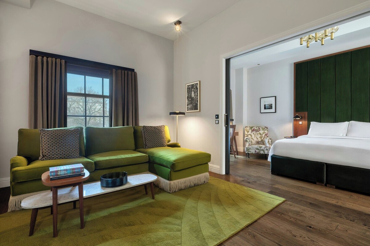 A room with a green sofa, a small table with books, a green rug, a bed with white bedding, and a door leading to another room.
