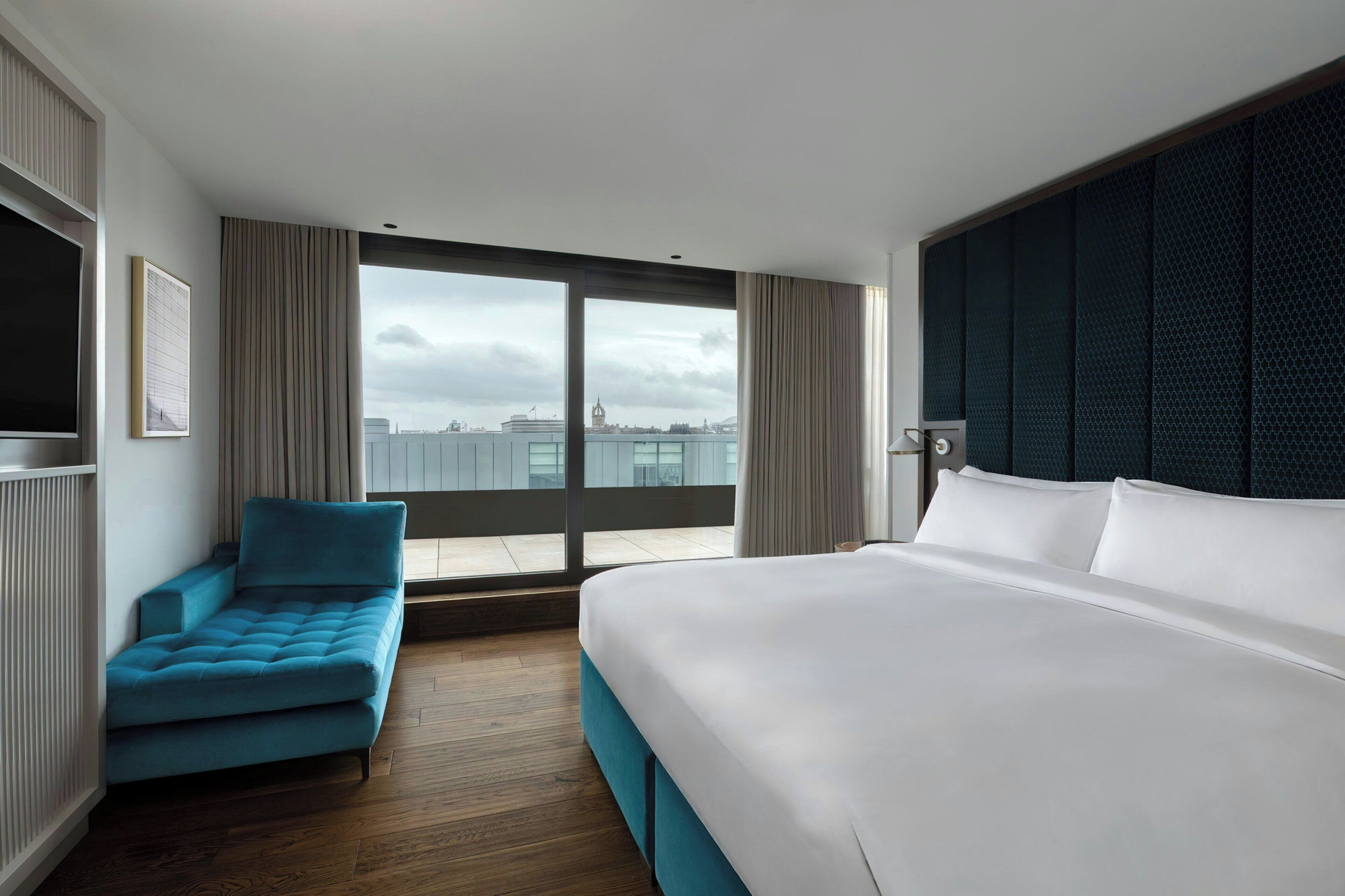 A hotel room with a large bed, a blue sofa, a view of the city, and a dark textured wall.