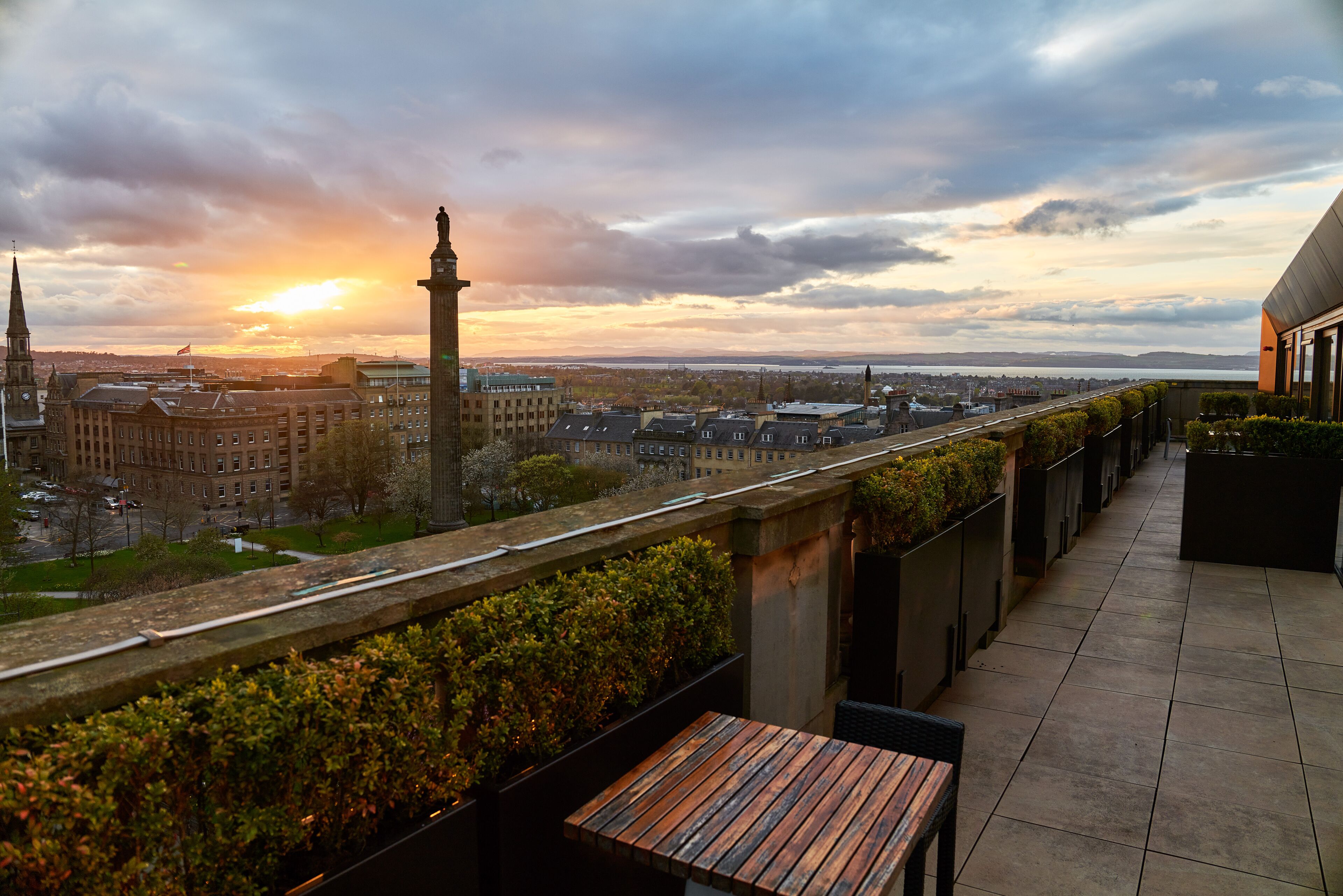 Ein Balkon mit Holztisch und -stühlen, mit Blick auf eine Stadtlandschaft mit Gebäuden und einem fernen Sonnenuntergang.